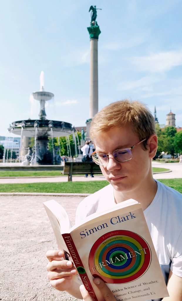 Aleksa Stankovic reading the book "Firmament" by Simon Clark in front of a statue and a fountain.
